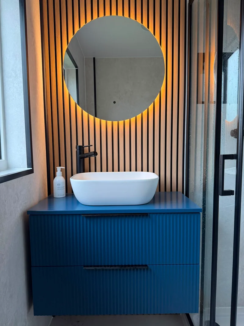 Blue fluted vanity with a countertop basin, backlit round mirror and wood-effect feature wall.