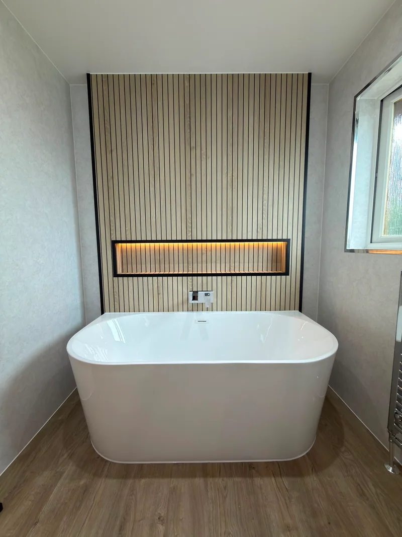White freestanding bath beneath an illuminated niche in a slatted feature wall.